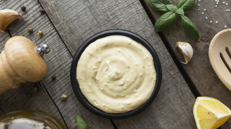 A homemade bowl of mayonnaise surrounded by basil leaves, garlic cloves, a lemon wedge, and ground pepper