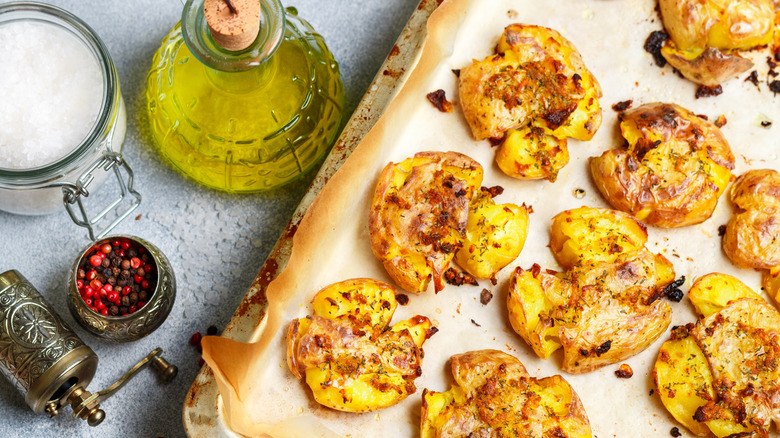 smashed potatoes on parchment-lined tray with oil, salt, and pink peppercorns nearby