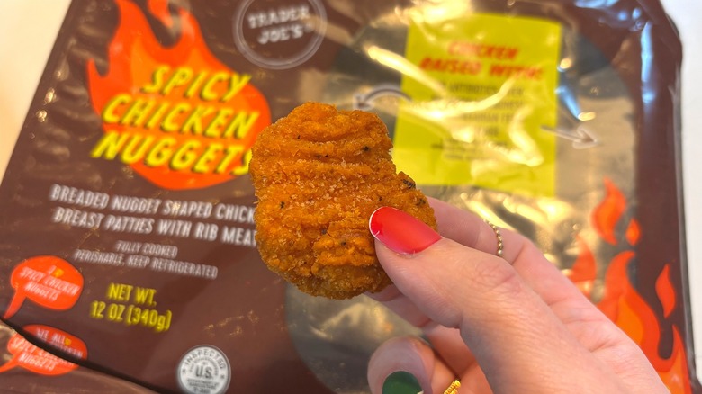 Hand holding a Trader Joe's spicy chicken nugget in front of package on white table