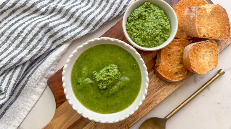 Bowl of spinach soup on wooden board with pesto and bread slices