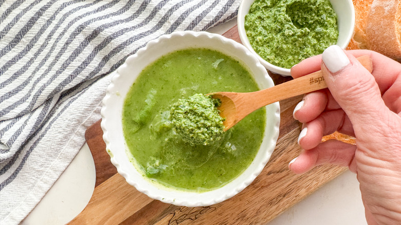 Hand spooning spinach pesto onto bowl of spinach soup