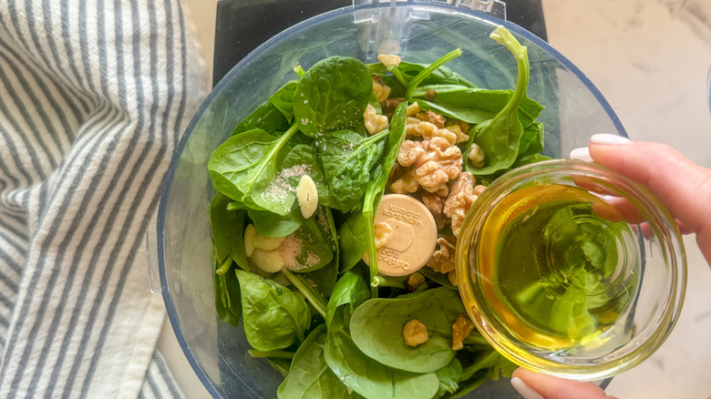 Hand pouring oil into food processor with spinach and other pesto ingredients