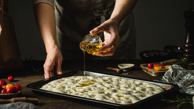 hands pouring olive oil over dimpled focaccia dough