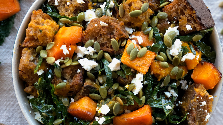 A bowl of kale panzanella salad, featuring squash cubes, croutons, seeds, kale, and cheese crumbles