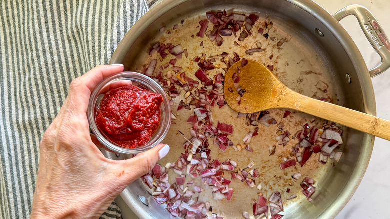 hand adding tomato paste to pan with onions