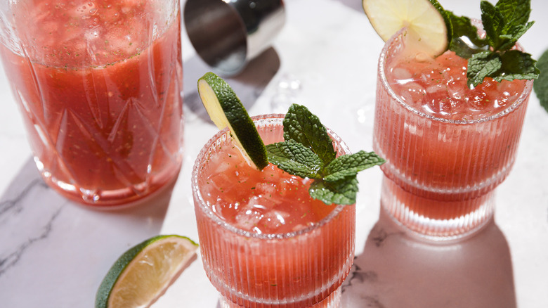 Two glasses and a pitcher with watermelon margarita mocktail, lime and mint around it