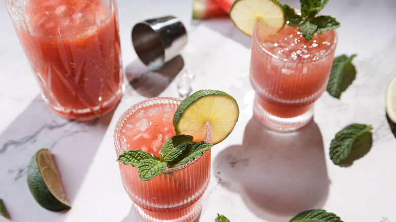 Two glasses and a pitcher with watermelon margarita mocktail, lime and mint around it