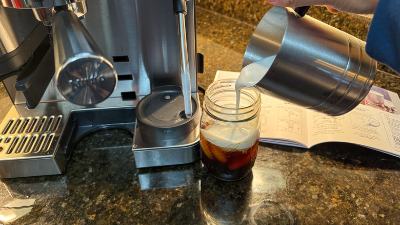 Person pouring cold foam on coffee with espresso machine in the background