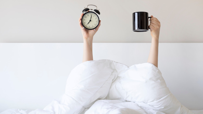 Arms holding up a cup of coffee and an alarm clock from bed