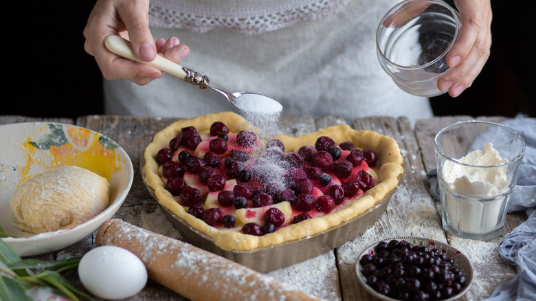 woman's hands sprinkling spoonful of granulated sugar over heart shaped pie topped with fresh blueberries and cranberries on flour dusted wooden table