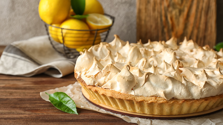 pastry crust tart toped with peaks of toasted meringue on wooden table with bowl of lemons in background