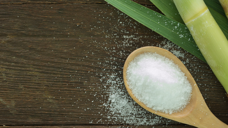 wooden spoon filled with granulated sugar next to raw sugarcane stalks and leaves on wooden table