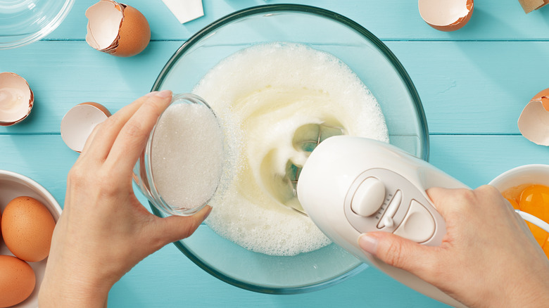 woman's hands pouring sugar into glass bowl while whipping egg whites with electric beater on blue table with raw eggs and eggshells scattered about