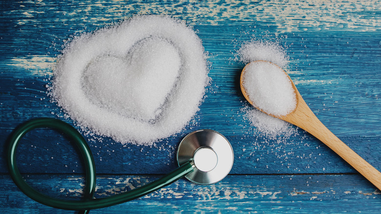 nutritional concept illustrated by spoonful of sugar, stethoscope, and heart shape drawn into a pile of sugar on a blue wooden table