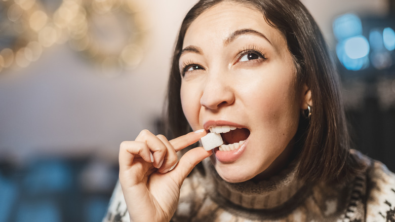 young brunette woman biting a cube of white sugar with blurred lights in background