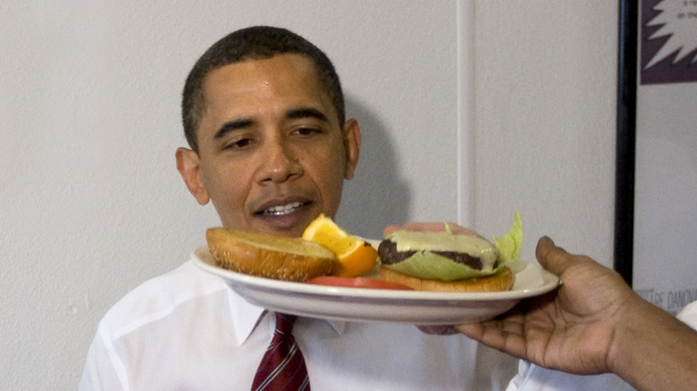 Barack Obama is served a cheeseburger on a white plate with an orange slice on the side