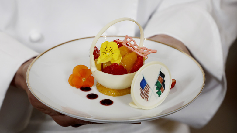 White House Pastry chef holds up a dessert created for a state dinner honoring Kenya with fresh raspberries, peaches, and edible candied flowers in an ornate white sugar basket.