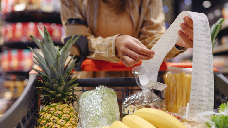 A young woman looks over her receipt from the grocery store while standing over her cart. Visible food includes pasta, pineapple, lettuce, and bananas.