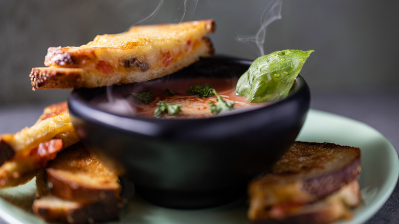 A golden brown toasted grilled cheese sandwich on a mint green plate with a steaming hot bowl of tomato basil soup, garnished with extra fresh green basil on top