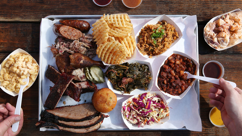 A tray of barbecue staples like meat, potatoes, beans, macaroni and cheese, coleslaw, and cornbread, placed on a white tray with various barbecue sauces on the side