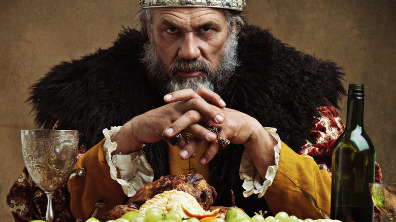 A medieval King sits in front of a large platter of food and wine