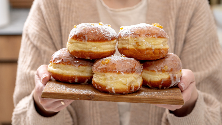 A person holds a wooden board with five glazed Polish paczki stacked on it.