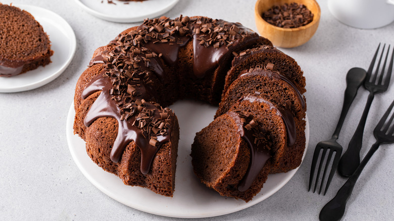 A sliced chocolate bundt cake sits on a table, surrounded by forks and small plates with slices.