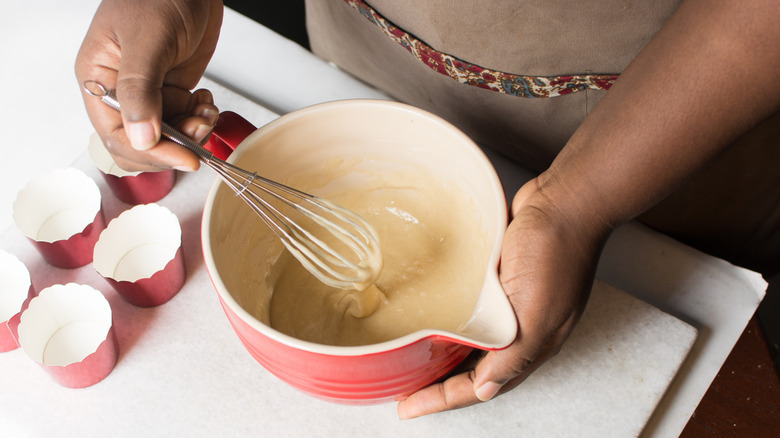 Someone's hands whisk a bowl of cake batter, while cupcake liners sit to the side.