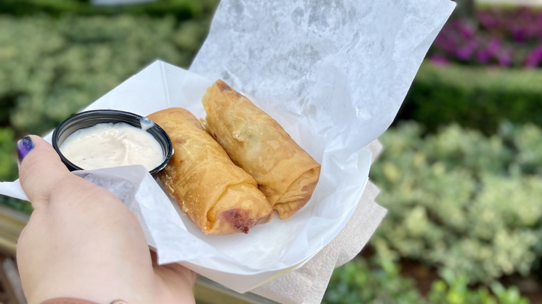 Hand holding a tray of two cheeseburger spring rolls with dipping sauce in small container