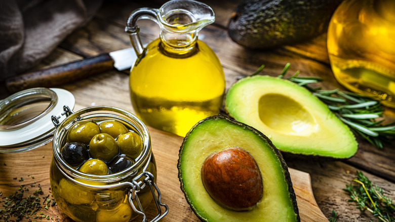 fresh avocad and avocado oil and whole olives next to olive oil on a wooden table