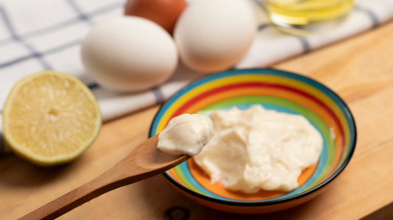 Wooden spoon with a portion of mayonnaise, below a colorful plate with mayonnaise next to a piece of lemon, a kitchen towel with three eggs and a glass with oil