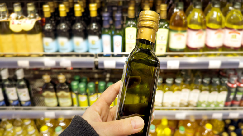 shopper's hand holding glass bottle of olive oil in front of a shelf full of bottles