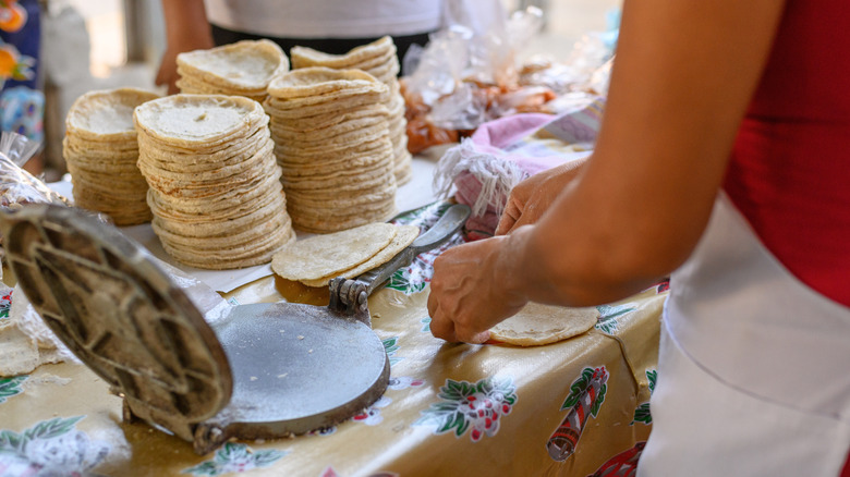 corn tortillas stacked next to a woman with a tortilla press