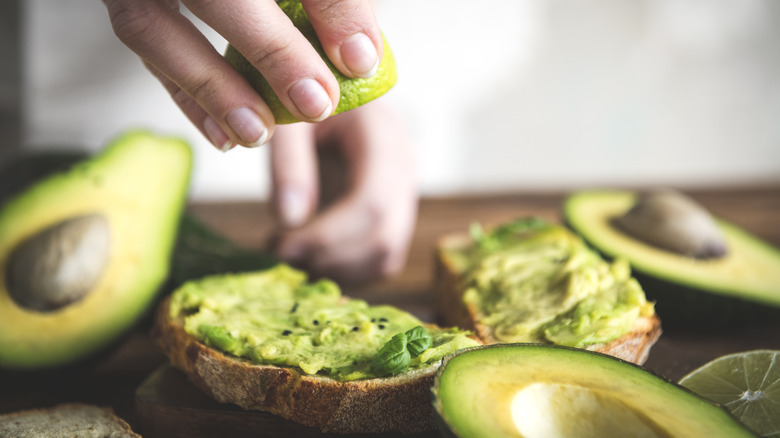 Hand squeezing lime over avocado toast next to ingredients