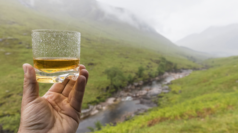 Hand holding a glass of scotch in front of a Scottish river