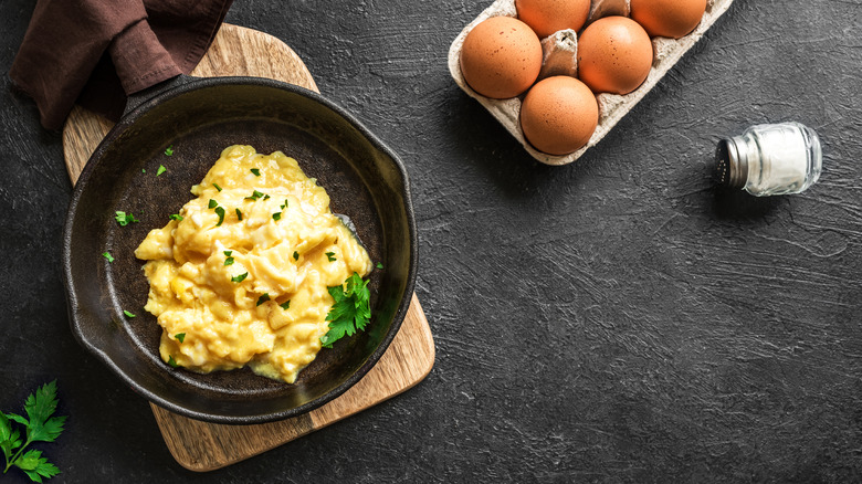 Scrambled eggs in a cast iron frying pan next to carton of eggs