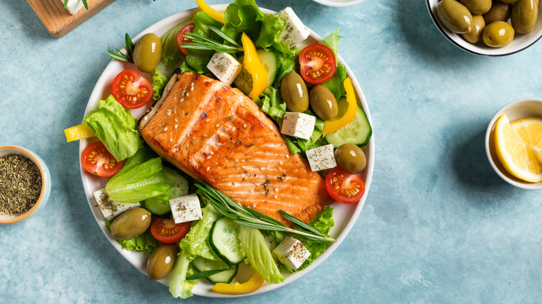 A piece of salmon on a bed of green lettuce, tomatoes, feta, peppers, olives, and cucumbers in a white bowl on a blue table surrounded by dishes of seasonings.