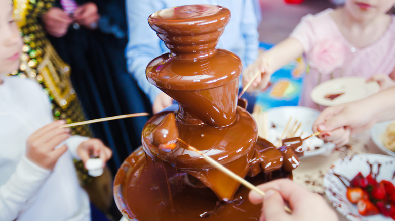 Several children gathered around a chocolate fountain dipping skewers of food into it