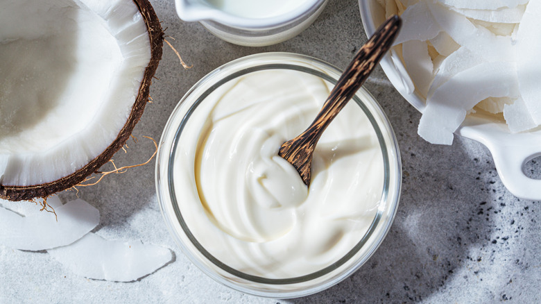 A jar of coconut yogurt with a wooden spoon inside. A halved coconut can be seen on the left and a bowl of coconut flakes on the right.